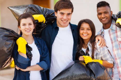 The image depicts four young adults standing outdoors, each holding black rubbish bags filled with waste. The individuals are smiling and appear to be engaged in a community or volunteer clean-up activity. They are dressed casually, with some wearing yellow gloves to handle the rubbish safely. The background shows a blurred outdoor environment, possibly a residential street or park, indicating a clean-up in progress. The scene is well-lit with natural daylight, emphasizing the communal effort and positive attitude towards rubbish removal. This visual aligns with services offered by Garden Clearance Putney, highlighting waste collection and rubbish removal in a local area, and the focus is on responsible disposal and maintaining a clean environment.