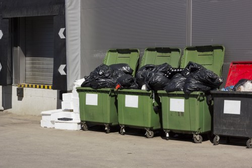 Four large wheelie bins are positioned in a row outside a commercial building, with three of them having green lids and one black. The contents visible through the partially open green lids appear to be tightly packed black rubbish bags, some tied with red ties, indicating general waste removal. To the right of the bins, a red container is also visible, likely used for waste disposal or collection. The area is paved, and the background features a grey metal wall with a partially visible grey roller shutter door, suggesting an industrial or commercial setting, possibly in Putney. A small stack of white bricks or tiles is placed on the ground beside the bins, and the scene is illuminated by daylight, emphasizing the neutral, utilitarian nature of the rubbish disposal area. This setup visually aligns with garden clearance services provided by Garden Clearance Putney, emphasizing waste collection and disposal for local properties.