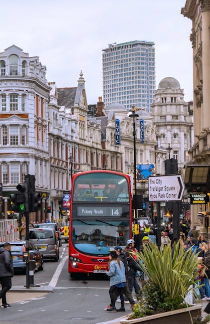 A busy urban street scene with a red double-decker bus displaying route number 14 and destination Putney Heath, surrounded by vehicles including cars and taxis, as well as pedestrians crossing and wal