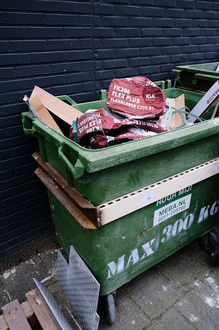 A green wheeled rubbish bin filled with red and black plastic bags, placed on a concrete pavement next to a dark brick wall. The bags appear to contain construction or garden waste, with visible text 