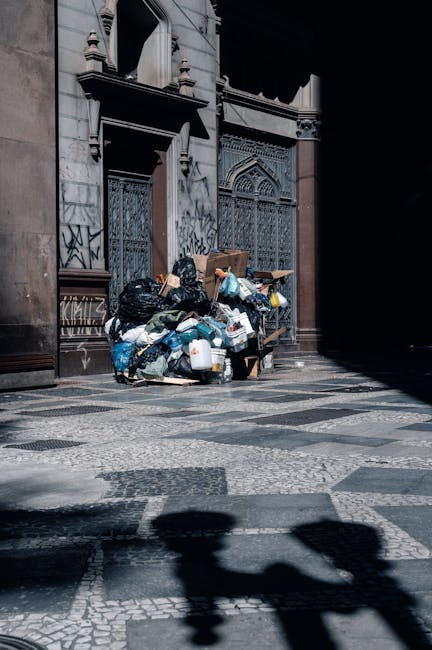 A green wheeled rubbish bin filled with red and black plastic bags, placed on a concrete pavement next to a dark brick wall. The bags appear to contain construction or garden waste, with visible text 